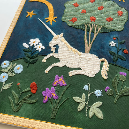Embroidered artwork of a unicorn in a garden with flowers and trees.