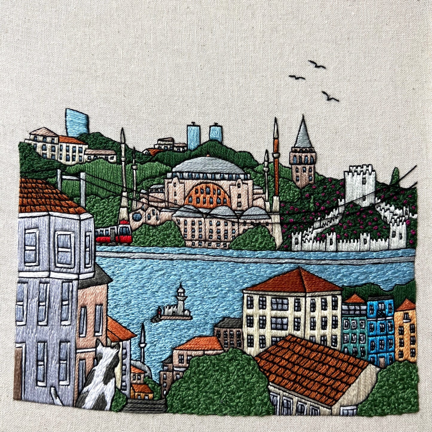 Embroidered cityscape with buildings, a river, and birds on a textured fabric background.