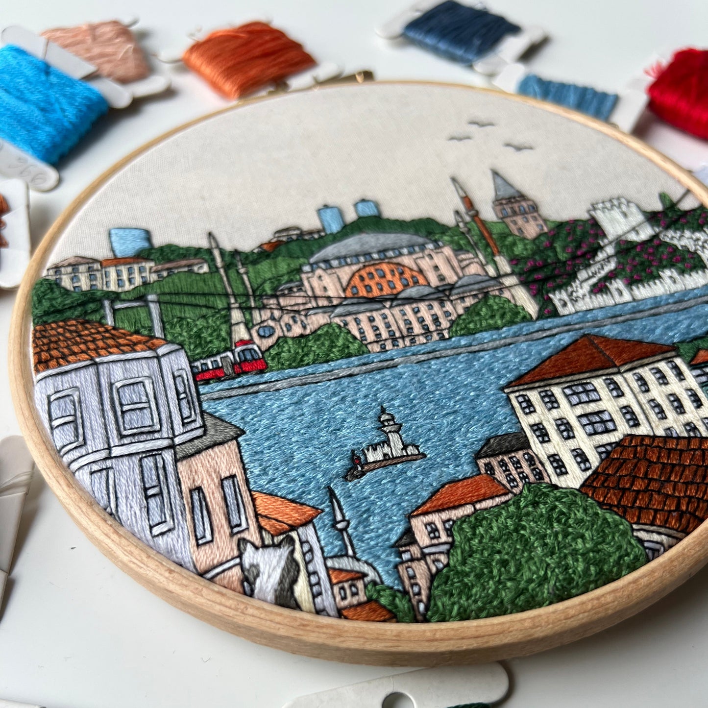Embroidery hoop with a colorful embroidered scene of a town by a river, surrounded by embroidery threads.