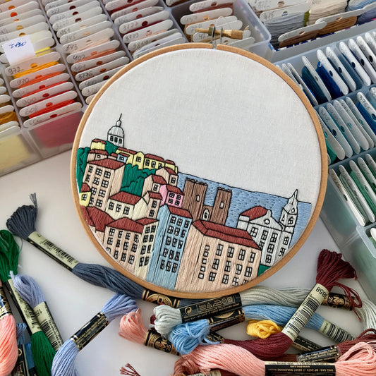 Embroidery hoop with a cityscape design surrounded by embroidery threads and supplies.
