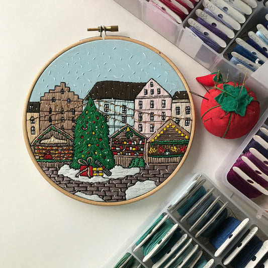 Embroidery hoop with a festive village scene surrounded by embroidery threads and tools.