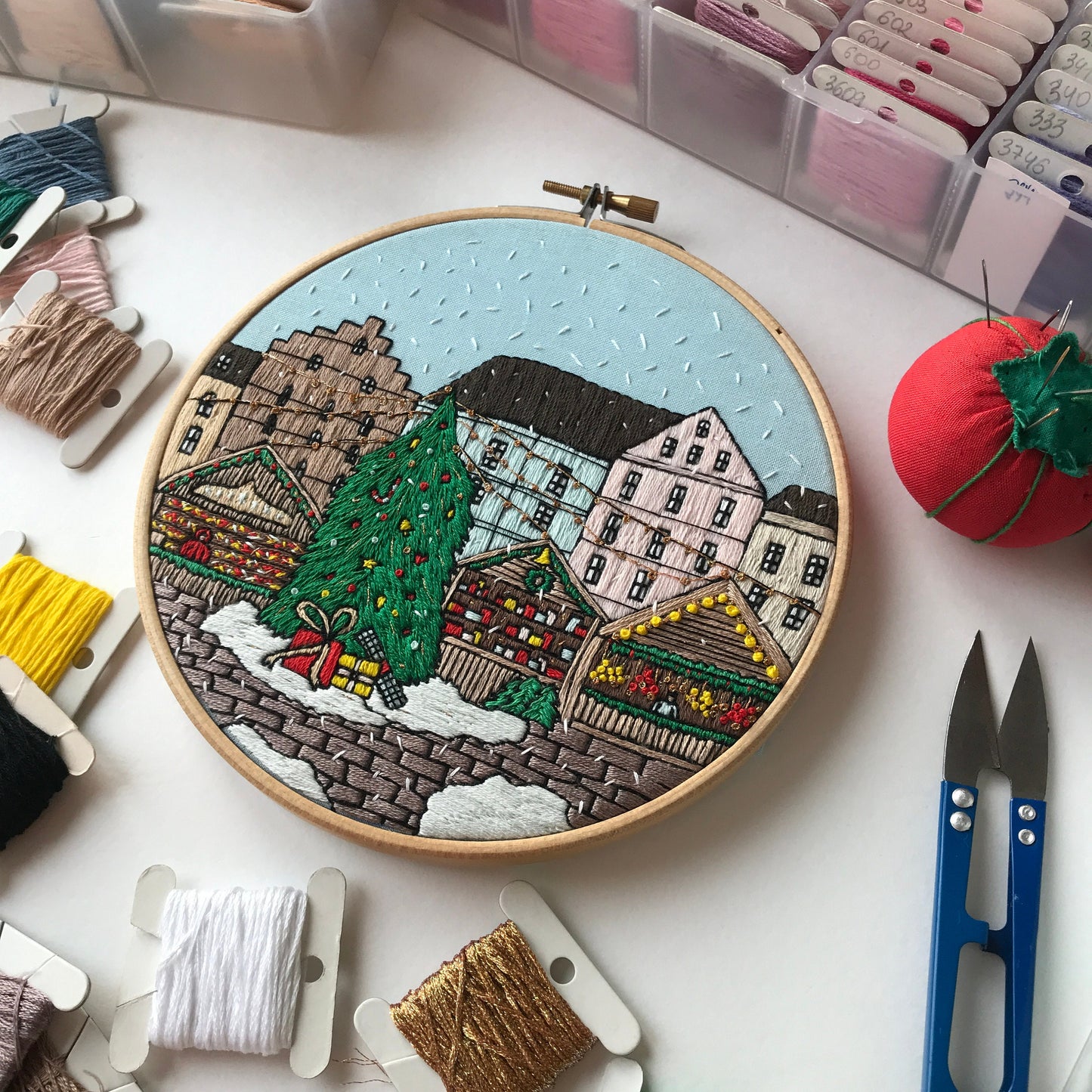 Embroidery hoop with a festive village scene surrounded by embroidery threads and tools on a white surface.
