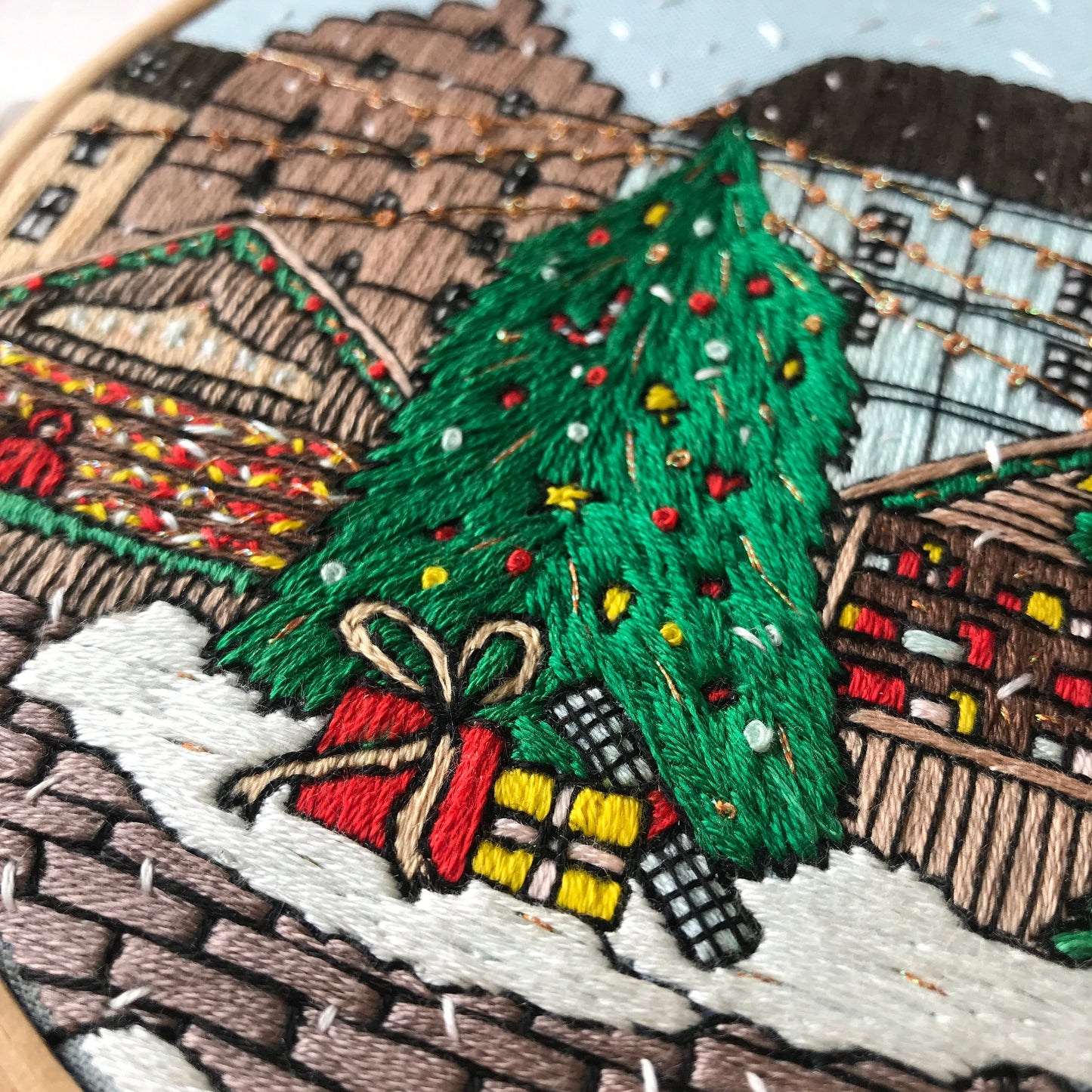 Embroidery of a Christmas tree with presents and lights on a snowy street scene.