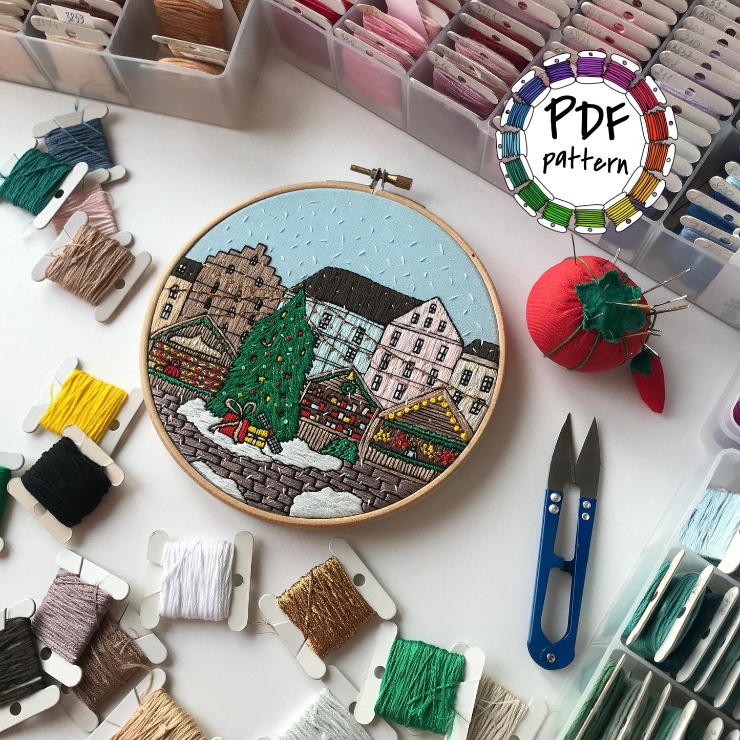 Embroidery hoop with a colorful cityscape design surrounded by embroidery threads and tools on a white surface.