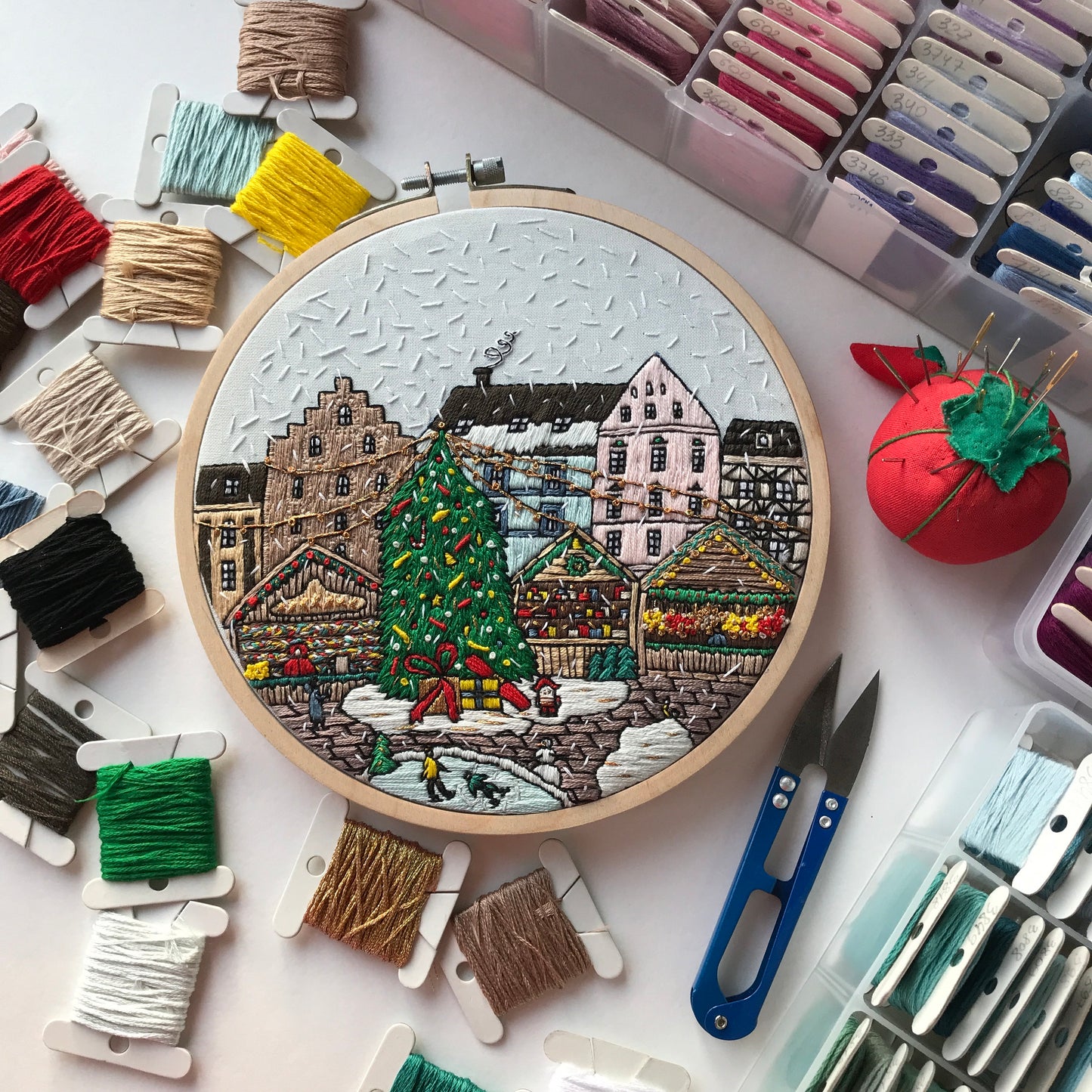 Embroidery hoop with a winter scene surrounded by embroidery threads and tools on a white surface