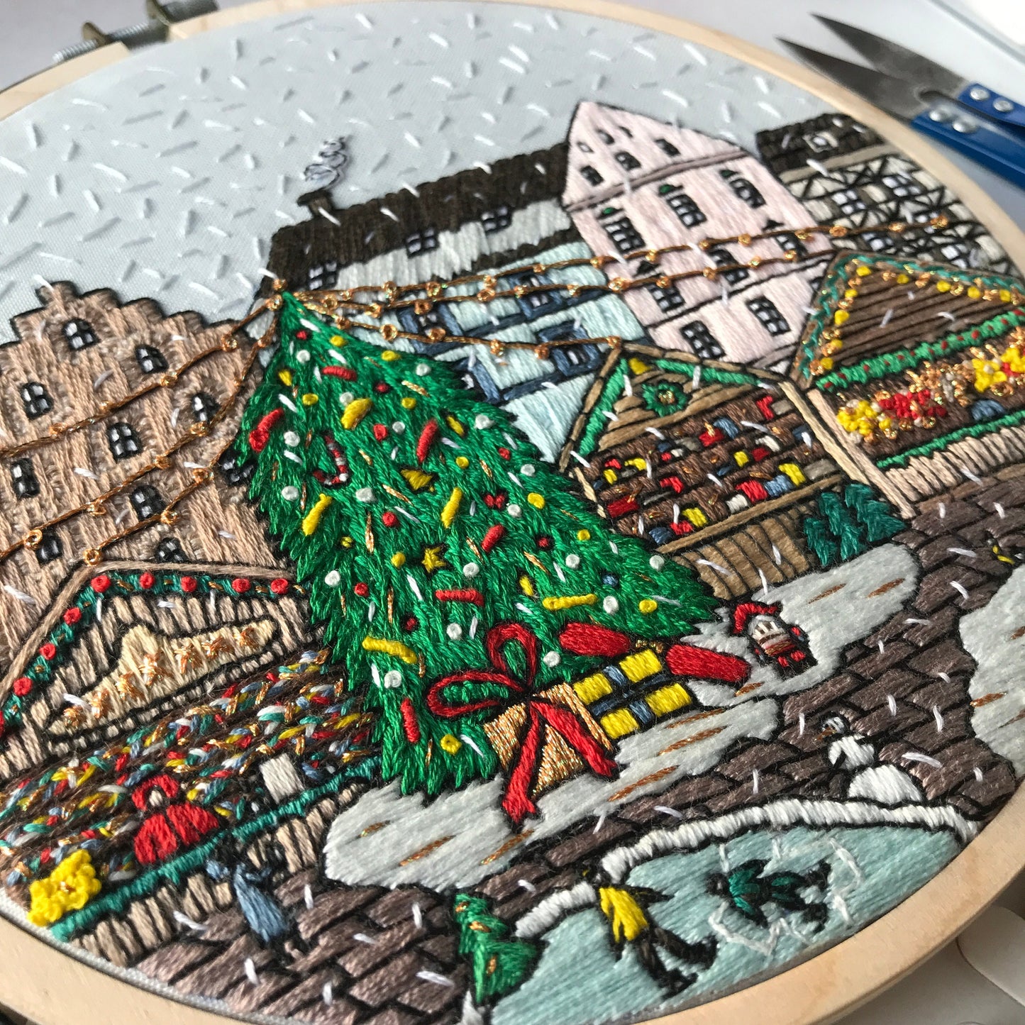 Embroidery of a festive scene with a Christmas tree and buildings in an embroidery hoop.