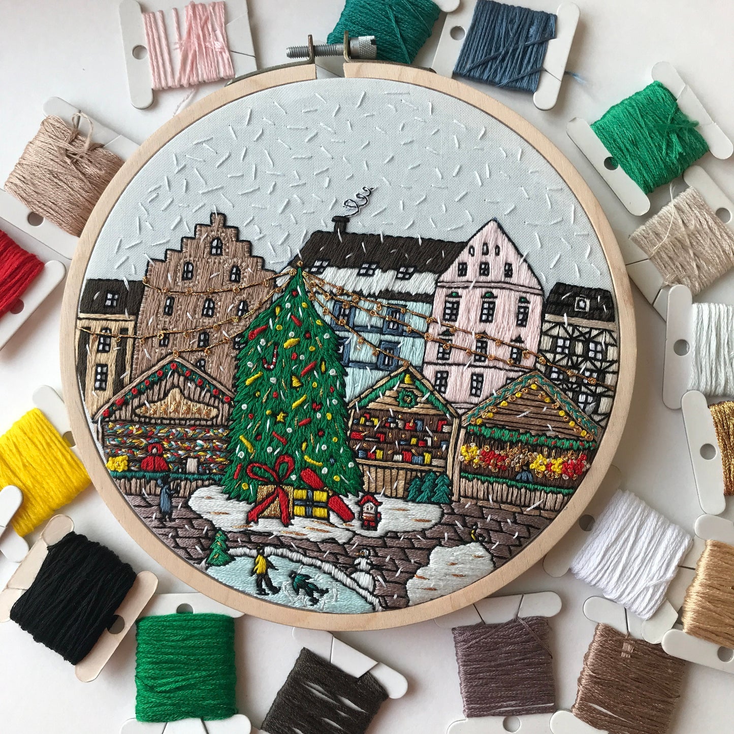 Embroidery hoop with a festive village scene surrounded by spools of thread.