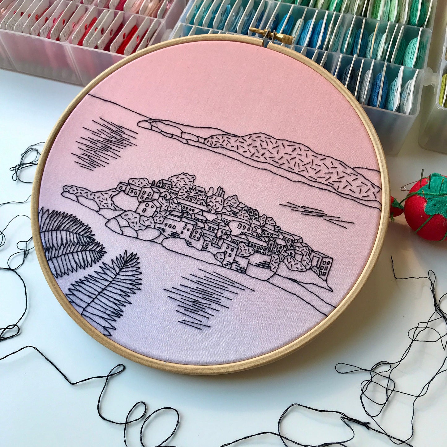 Embroidery hoop with a pink background and black line art of a village.