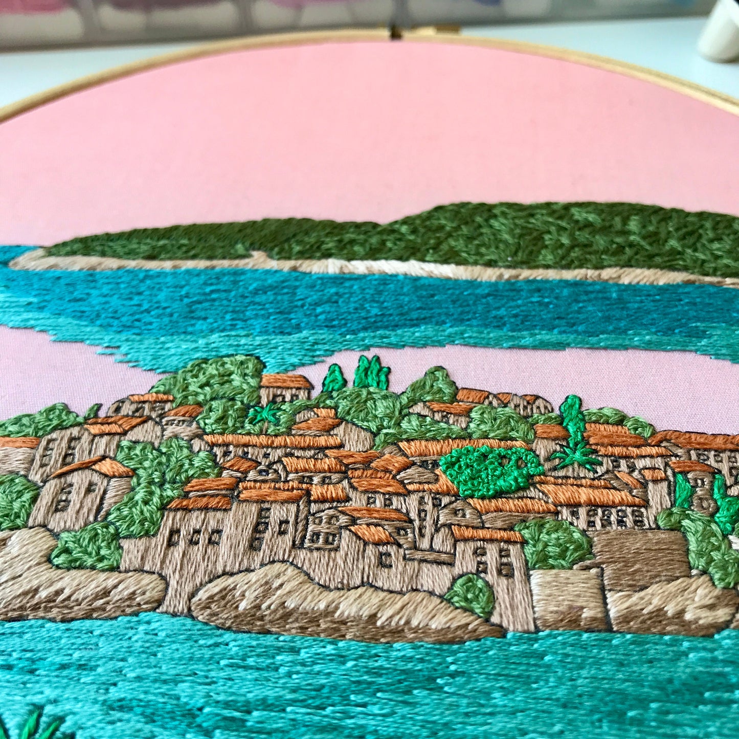 Embroidery of a coastal scene with buildings, trees, and water.