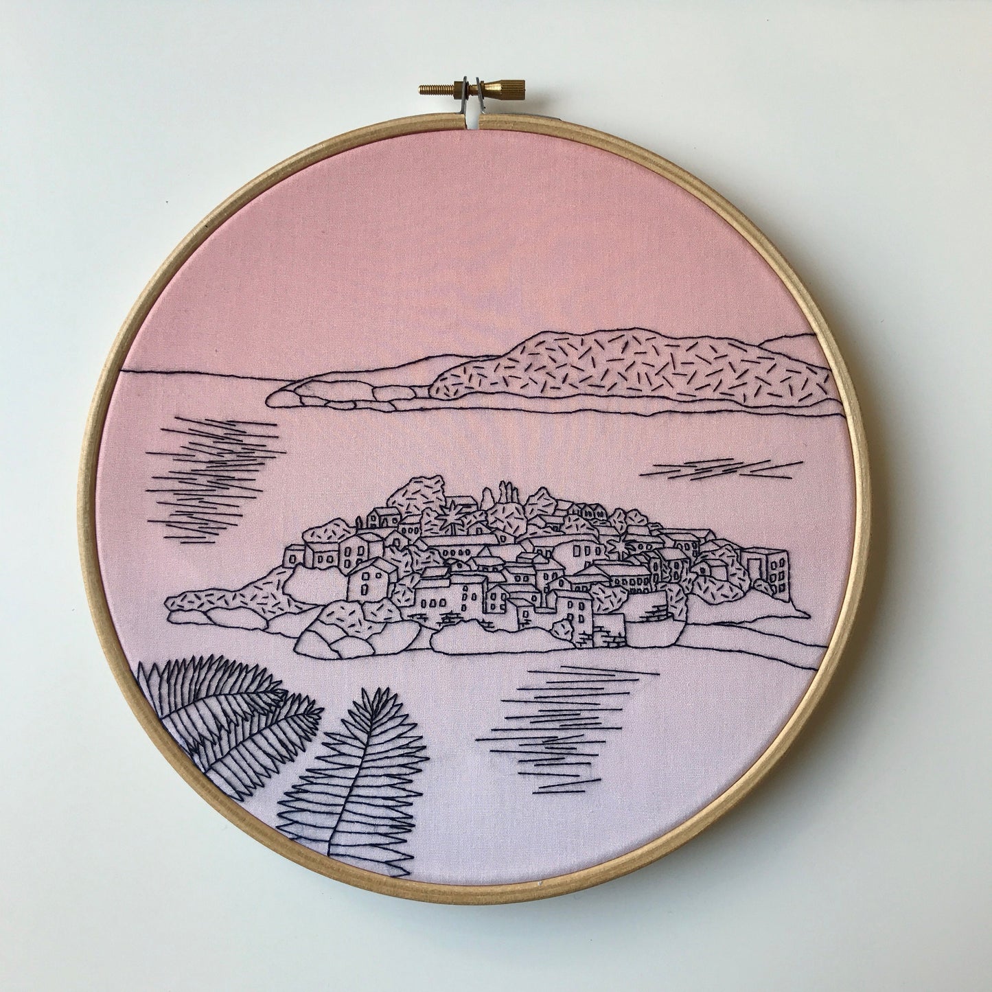 Embroidery hoop with a pink background and black line art of a coastal town.