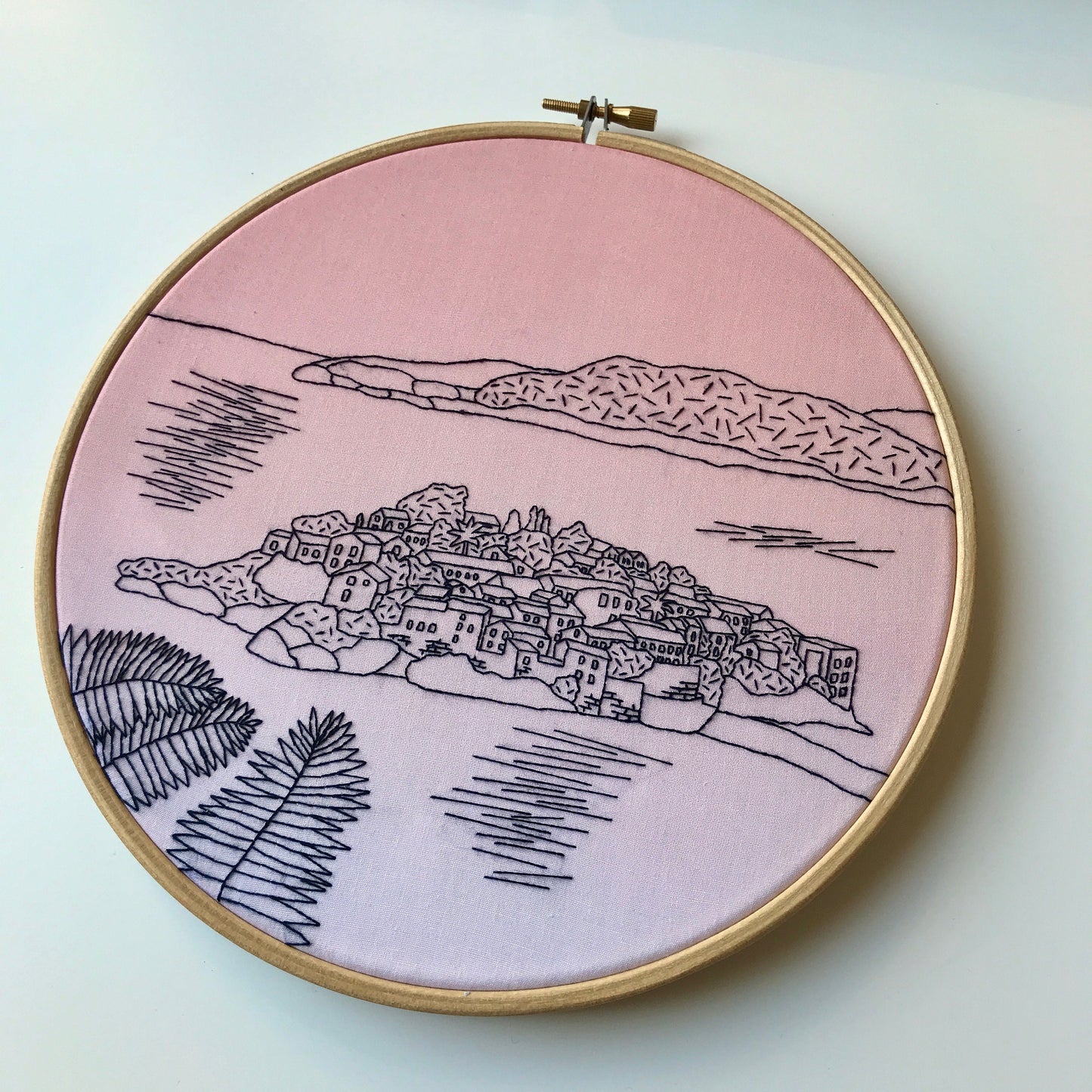 Embroidery hoop with pink fabric and black embroidered design of a coastal village.