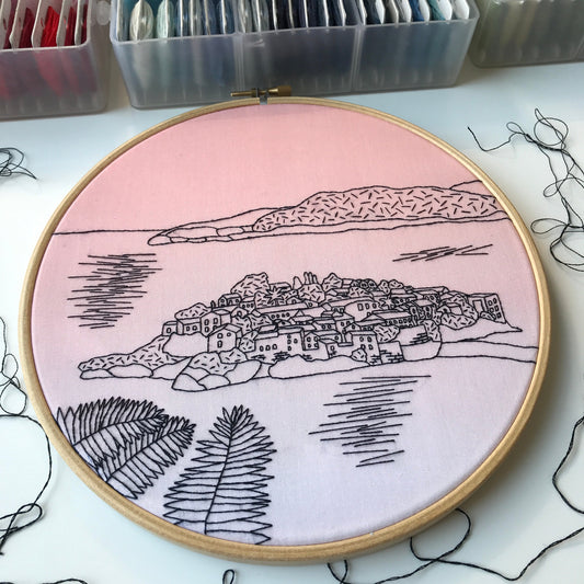 Embroidery hoop with a landscape design on a pink background, surrounded by embroidery supplies.