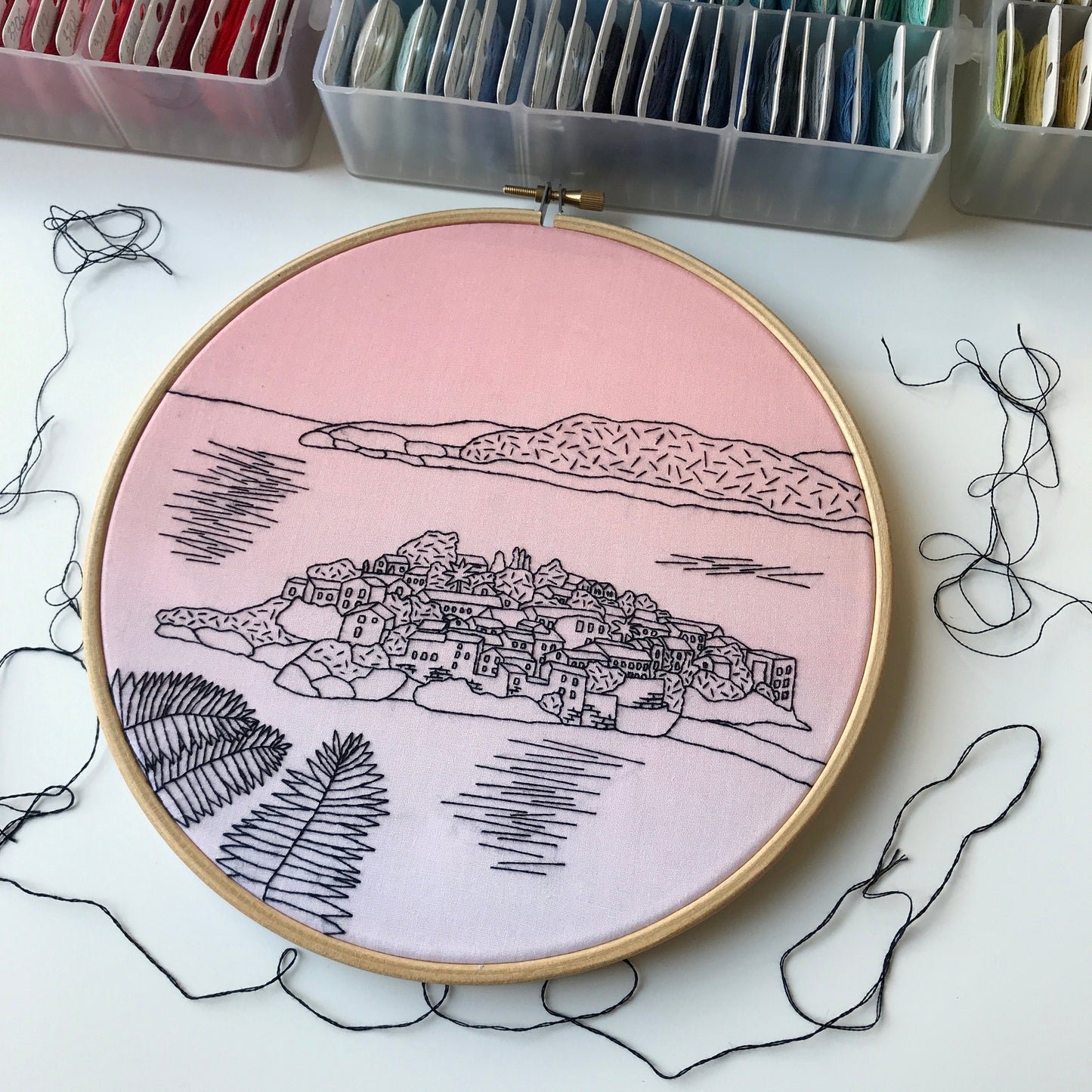Embroidery hoop with a pink background and black line art of a village, surrounded by embroidery supplies.