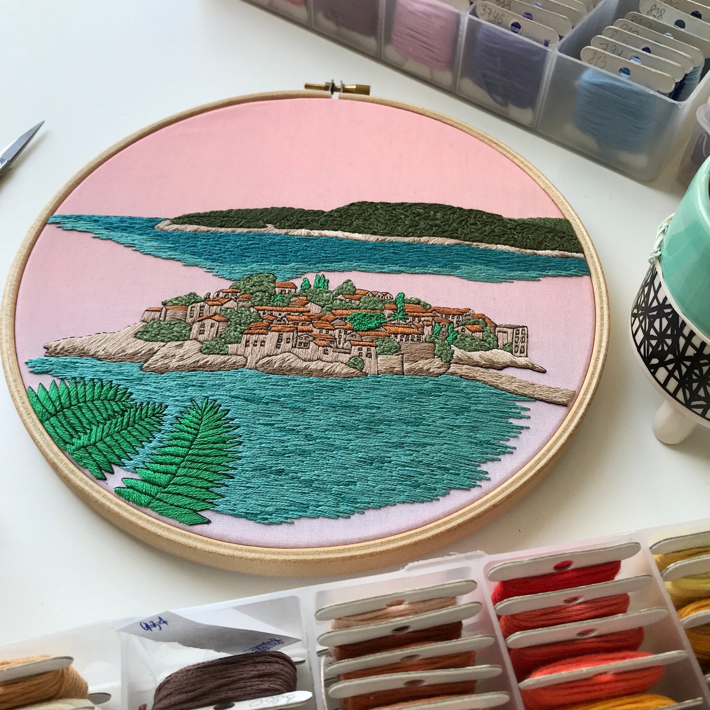 Embroidery hoop with a scenic landscape design on a table with embroidery supplies.
