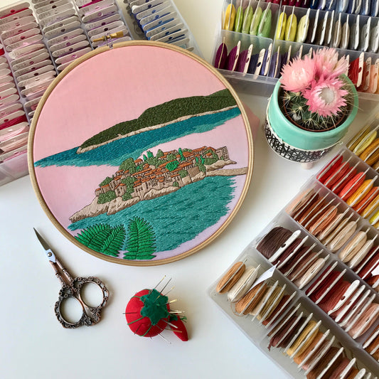 Embroidery hoop with a colorful landscape design on a table with embroidery supplies.