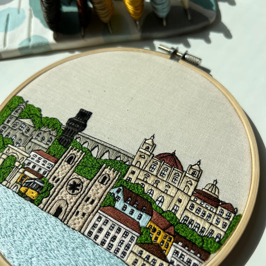 Embroidery hoop with a cityscape design on fabric, with thread spools in the background.