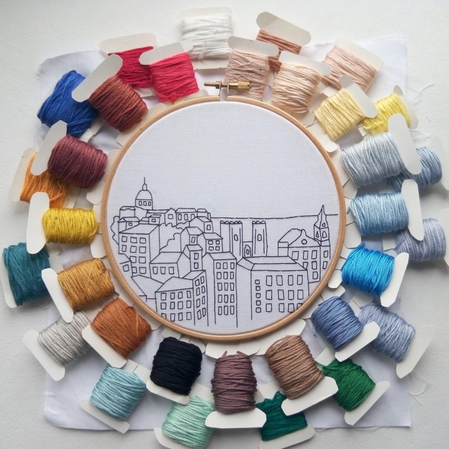 Embroidery hoop with cityscape design surrounded by colorful thread spools on a light background