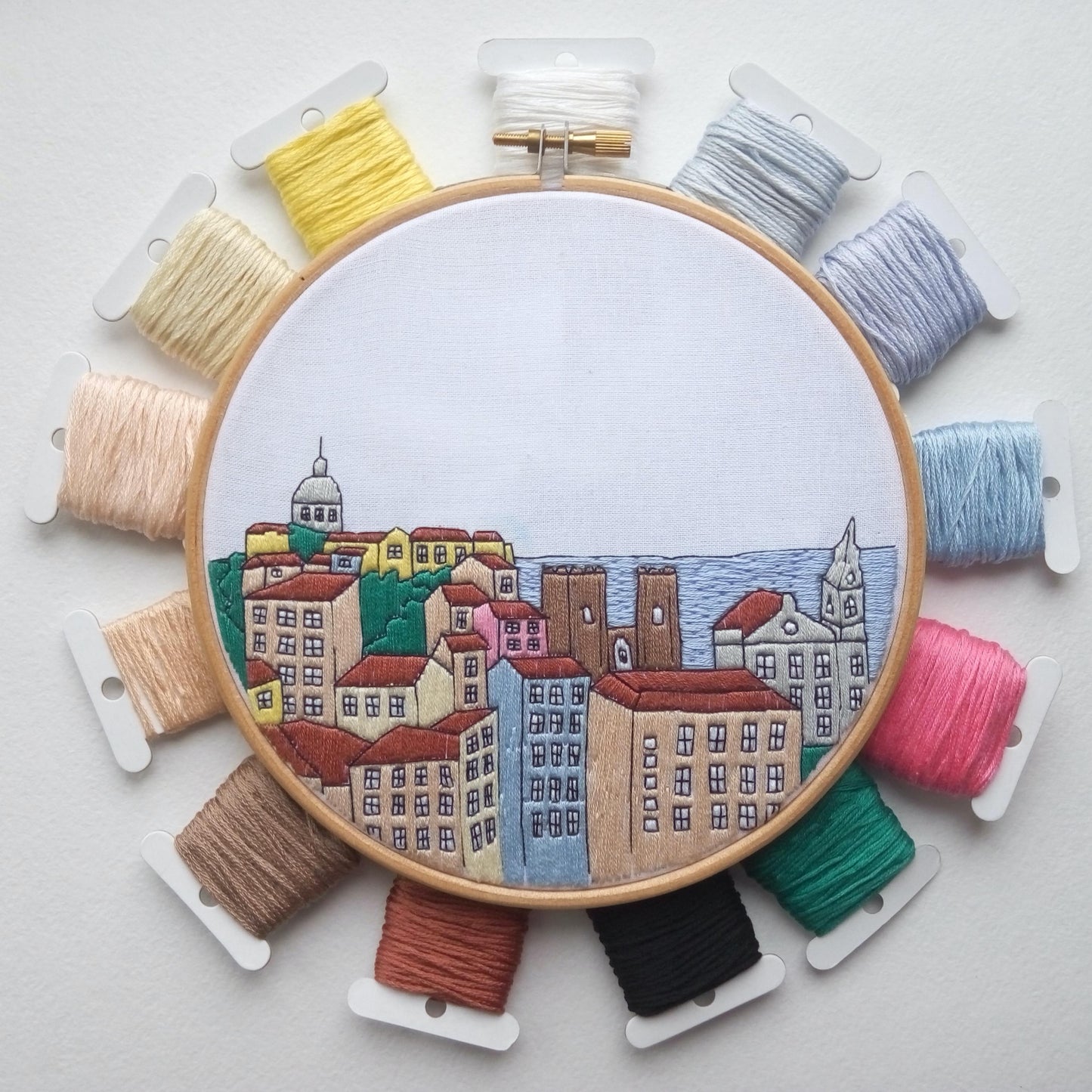 Embroidery hoop with a colorful cityscape design surrounded by bobbins of thread on a light gray background