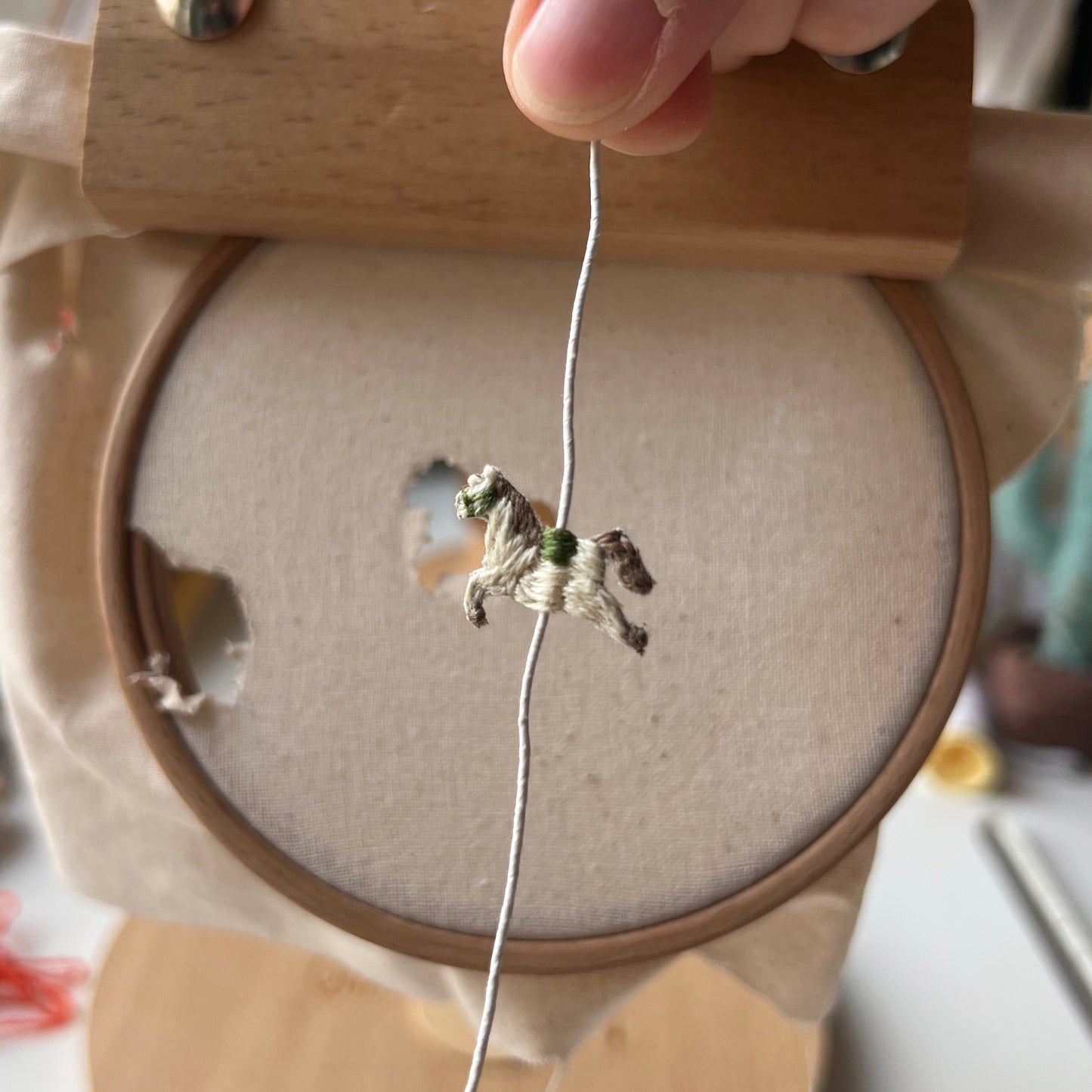 Small embroidered horse on a wire held by a hand against a wooden hoop 