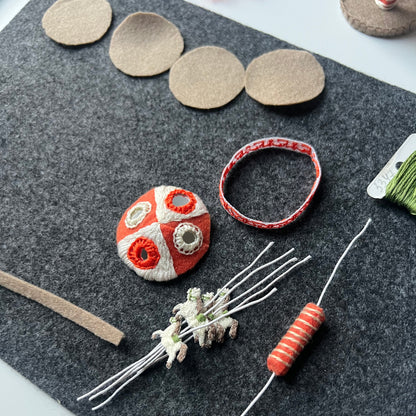 Craft materials include an Aida strip with embroidery, circular pieces of felt, and decorative elements on a gray felt.