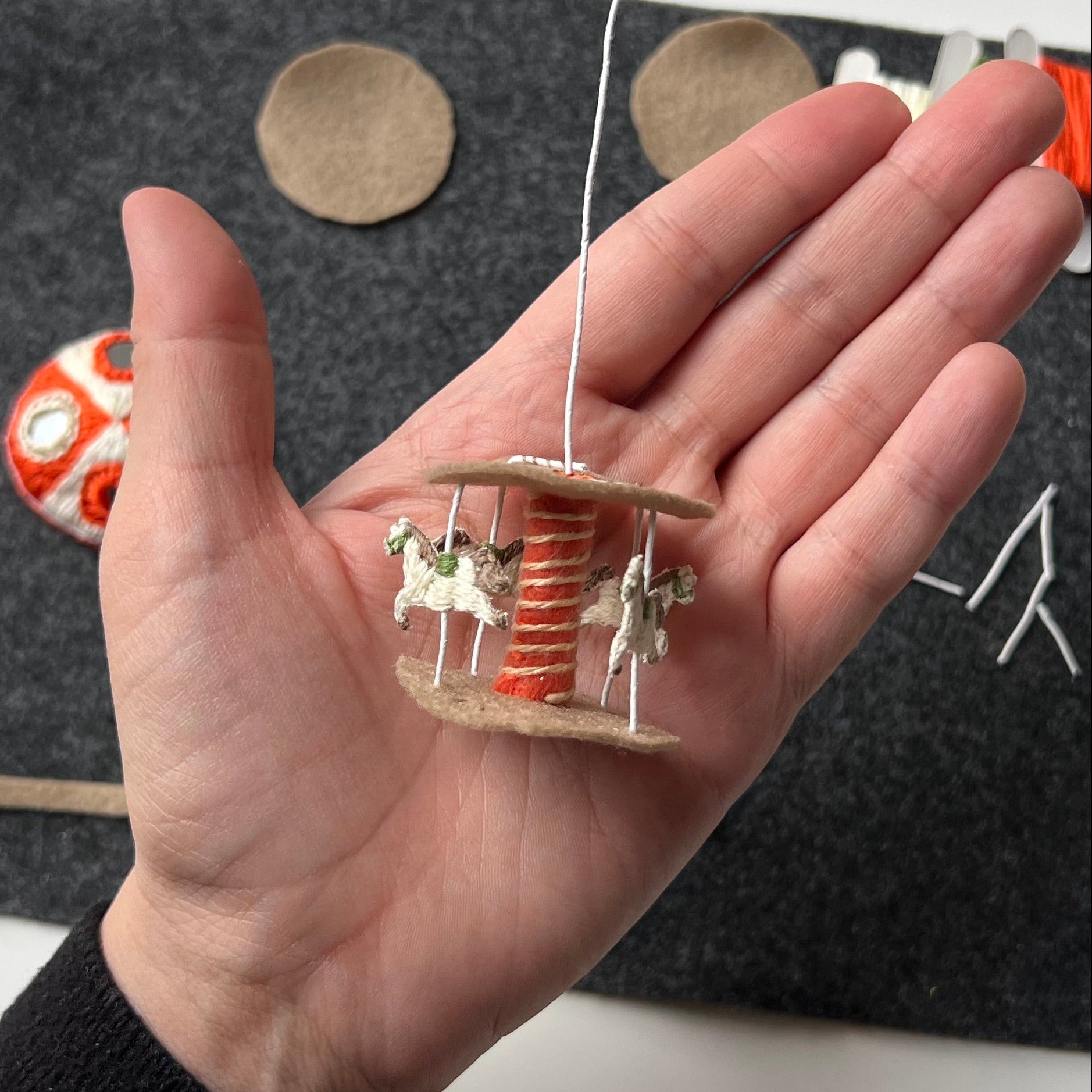 Hand holding a miniature stumpwork carousel in progress with horses on a dark surface
