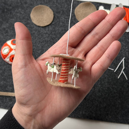 Hand holding a miniature stumpwork carousel in progress with horses on a dark surface
