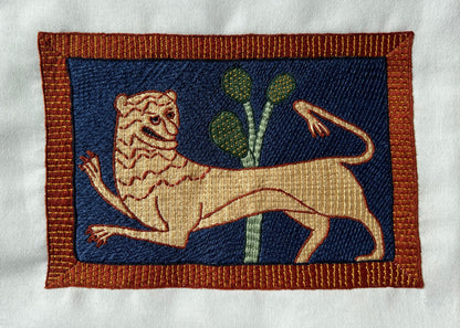 Embroidered patch of a lion with plants on a blue background, framed by brown border.
