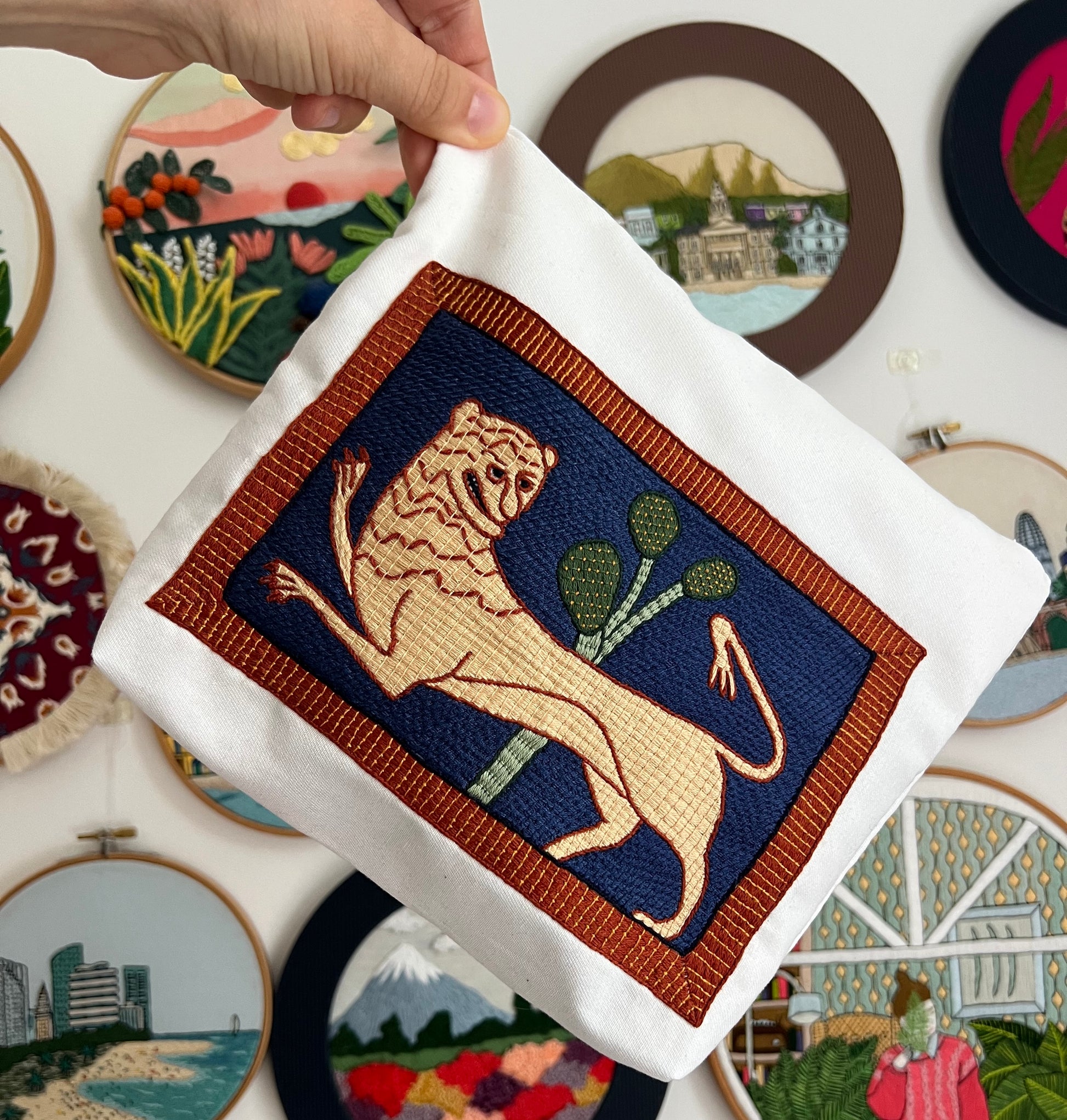 Embroidered pouch with a lion design held by a hand, surrounded by other embroidered pieces on a white background.