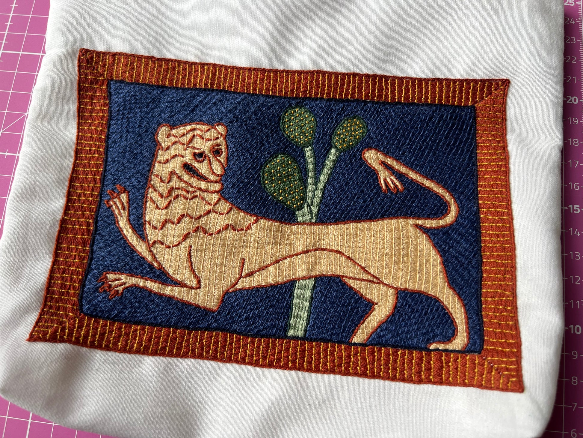 Embroidered patch of a lion and a plant on a blue background with brown border, on a white fabric.