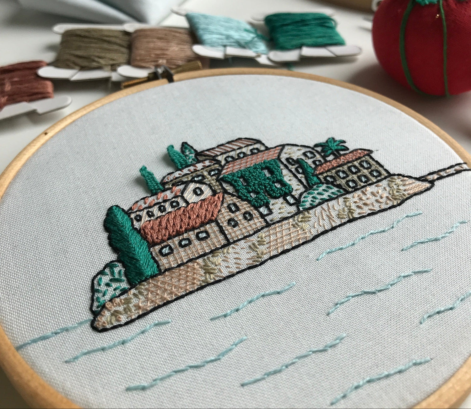 Embroidery hoop with a colorful embroidered scene of buildings and water on a light background.