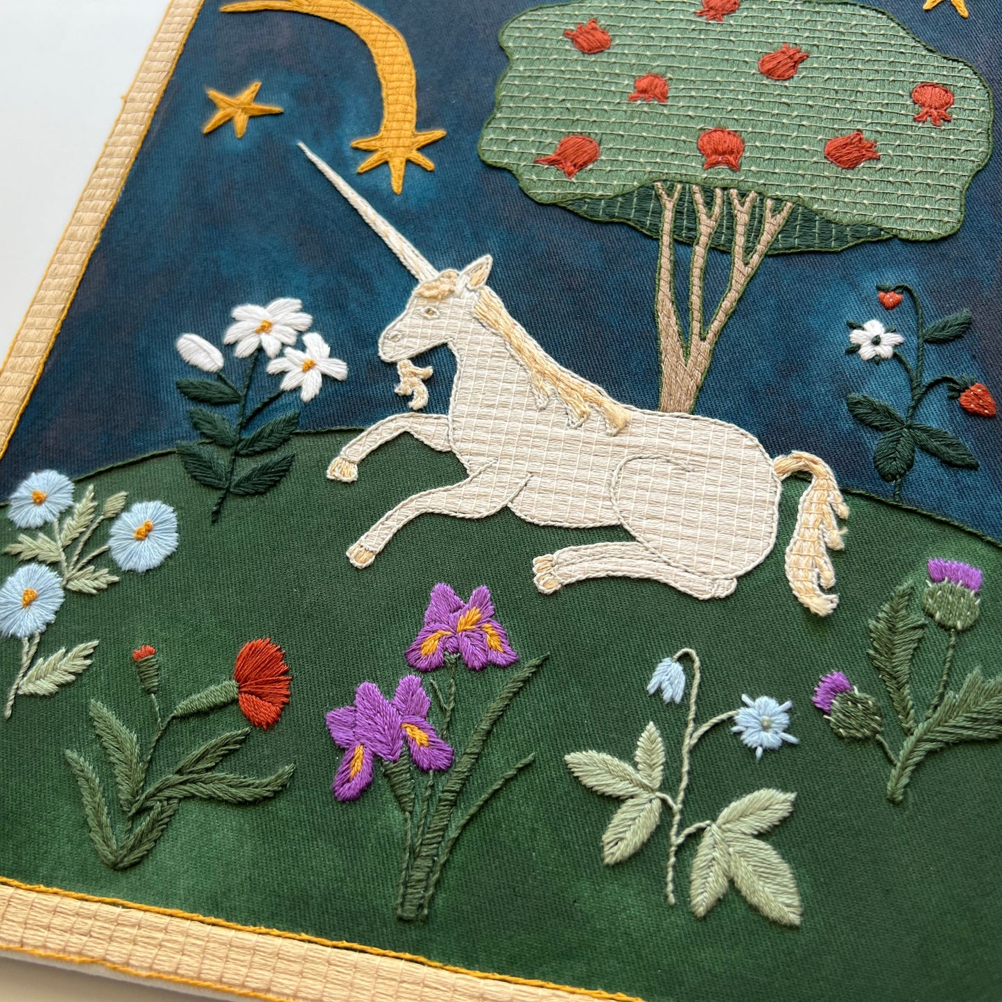 Embroidered artwork of a unicorn in a garden with flowers and trees.