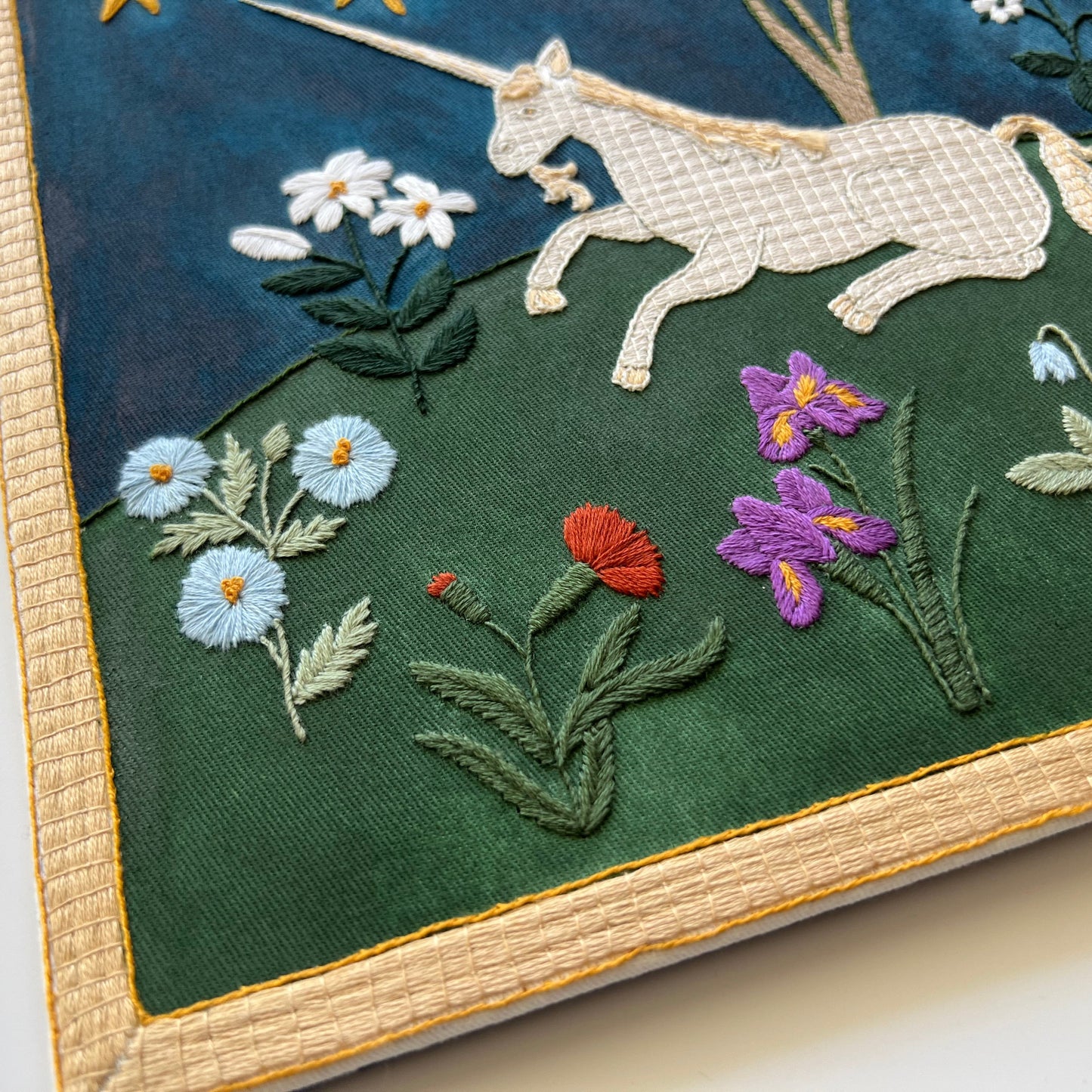 Embroidered artwork of a unicorn and flowers on a dark green fabric background.