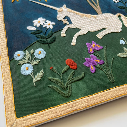 Embroidered artwork of a unicorn and flowers on a dark green fabric background.