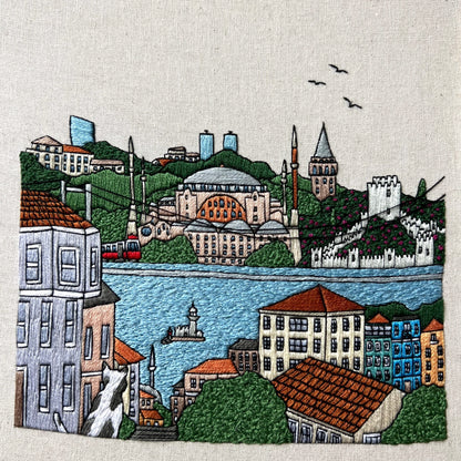 Embroidered cityscape with buildings, a river, and birds on a textured fabric background.