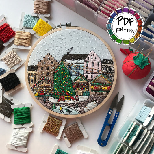 Embroidery hoop with a festive village scene surrounded by embroidery threads and tools on a white background.