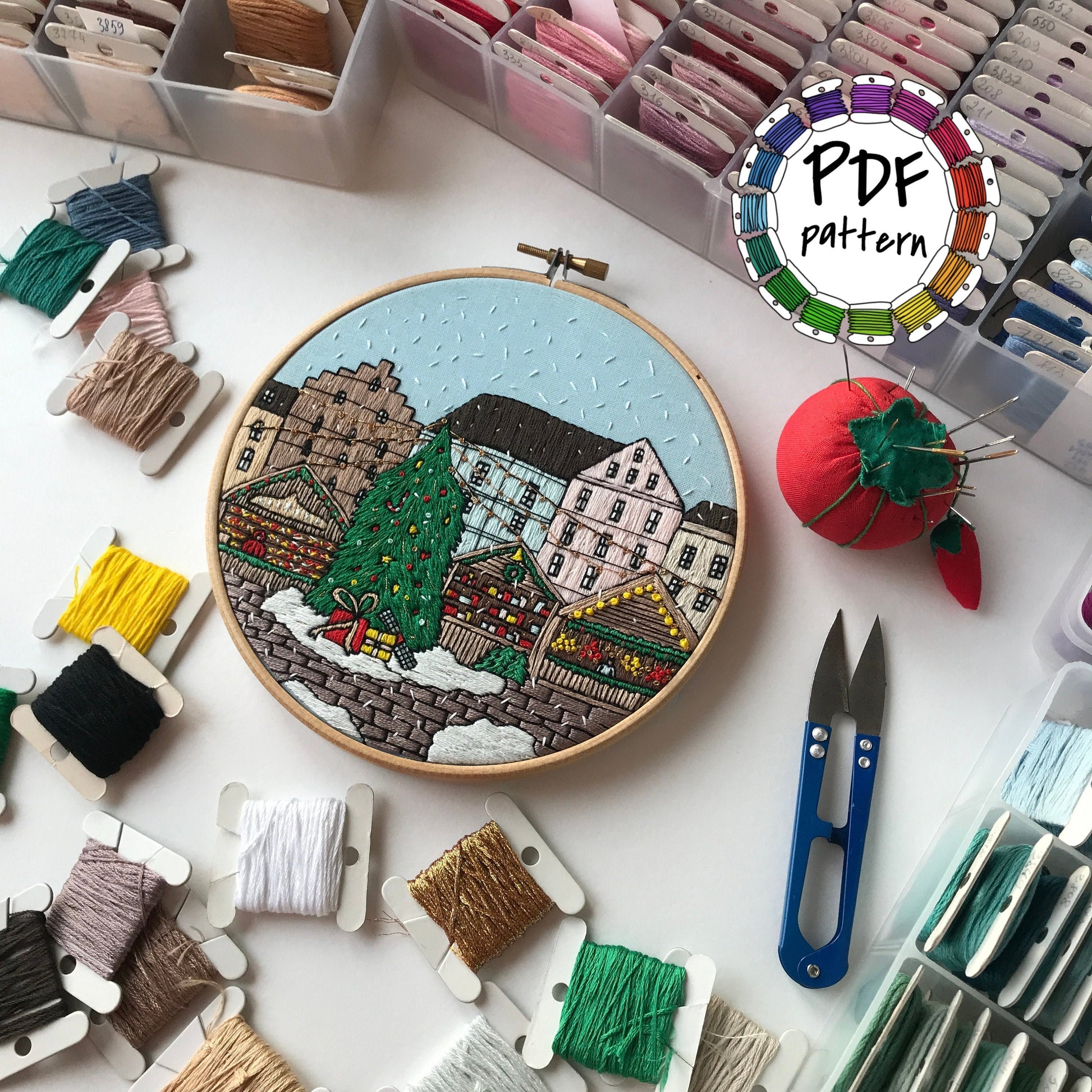 Embroidery hoop with a colorful cityscape design surrounded by embroidery threads and tools on a white surface.