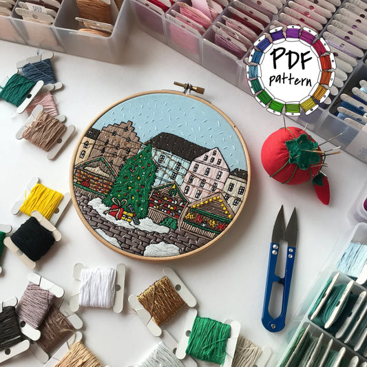 Embroidery hoop with a colorful cityscape design surrounded by embroidery threads and tools on a white surface.