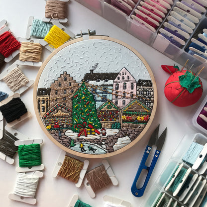 Embroidery hoop with a winter scene surrounded by embroidery threads and tools on a white surface