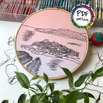 Embroidery hoop with a scenic design on a pink background, surrounded by embroidery supplies.