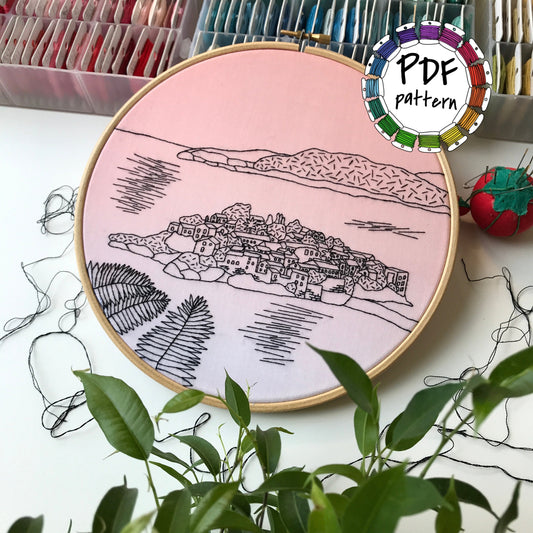 Embroidery hoop with a scenic design on a pink background, surrounded by embroidery supplies.