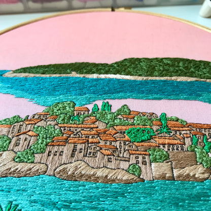 Embroidery of a coastal scene with buildings, trees, and water.