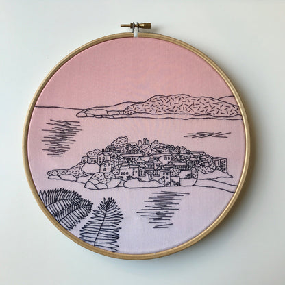 Embroidery hoop with a pink background and black line art of a coastal town.