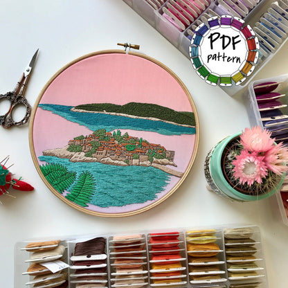 Embroidery hoop with island design, embroidery threads, scissors, and a cactus on a white surface.
