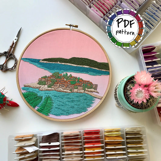 Embroidery hoop with island design, embroidery threads, scissors, and a cactus on a white surface.