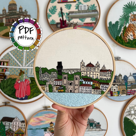 Embroidery hoop with a colorful Lisbon cityscape design held by a hand, surrounded by other embroidered scenes.
